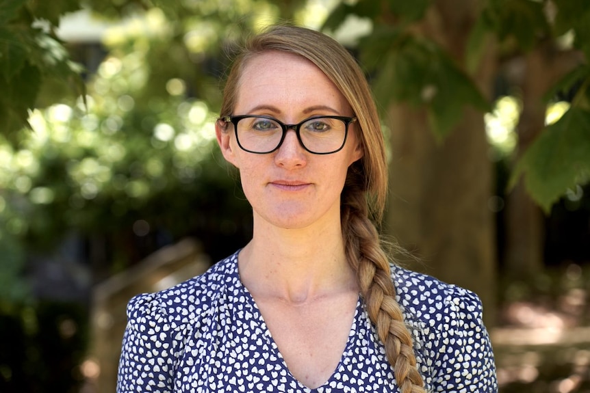 A lady with a long plait stands in front of a tree with a serious expression