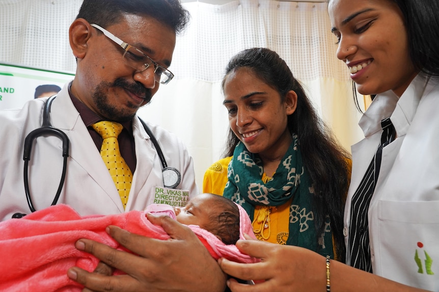 Three adults stand around a small baby girl. 