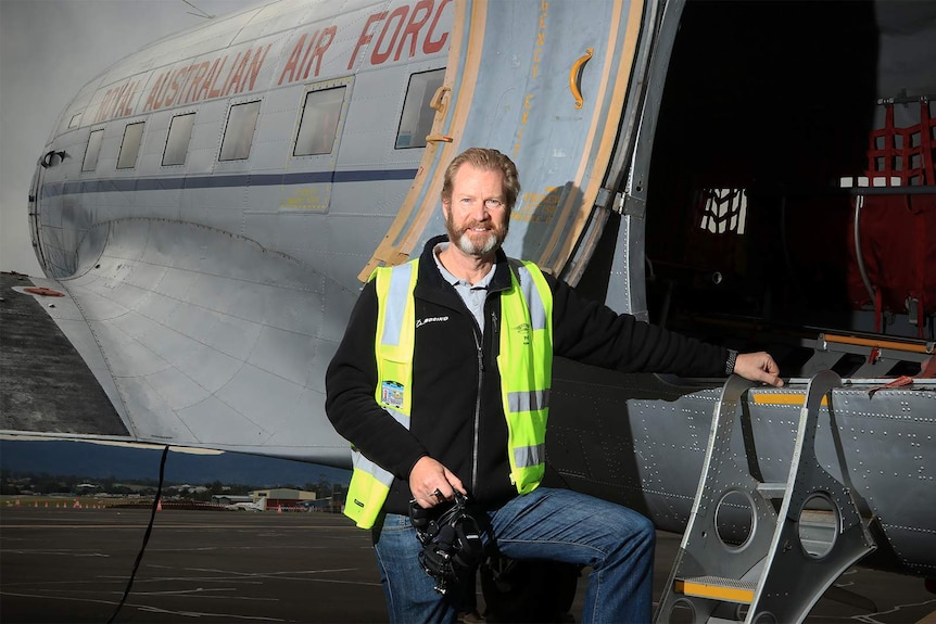 A bearded man of late middle age stands with one leg on a step on a DC-3 aircraft, wearing a high-vis vest.