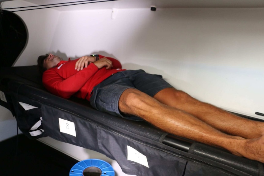 A man lying down in a cramped space to sleep on the boat.
