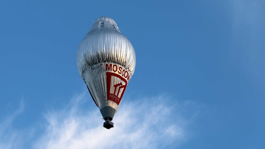 A large silver hot air balloon sits in a pale blue sky, with a wispy cloud below.