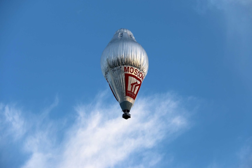 A large silver hot air balloon sits in a pale blue sky, with a wispy cloud below.