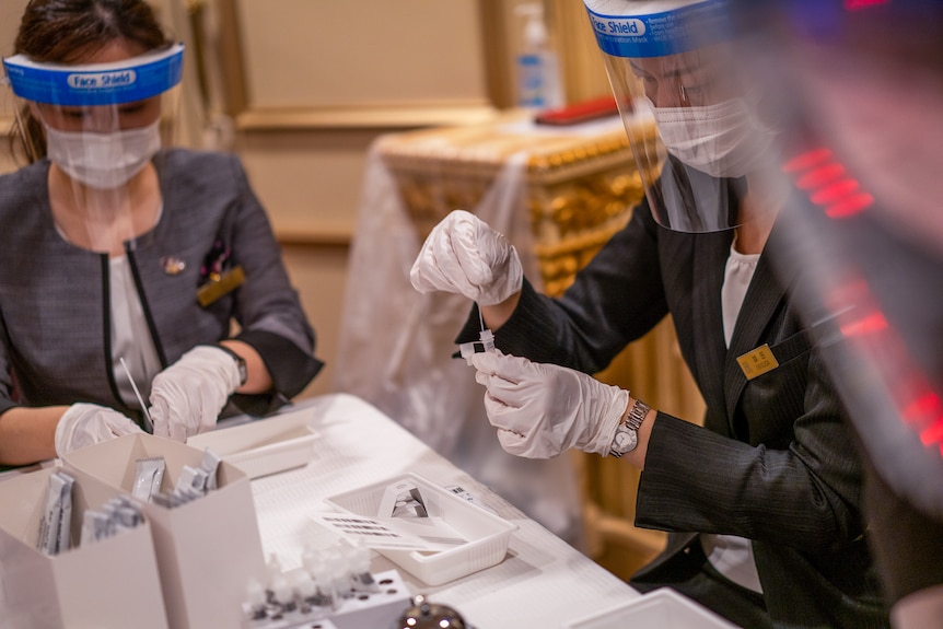 Two women in masks and shields put nasal swabs in testing vials in a hotel lobby