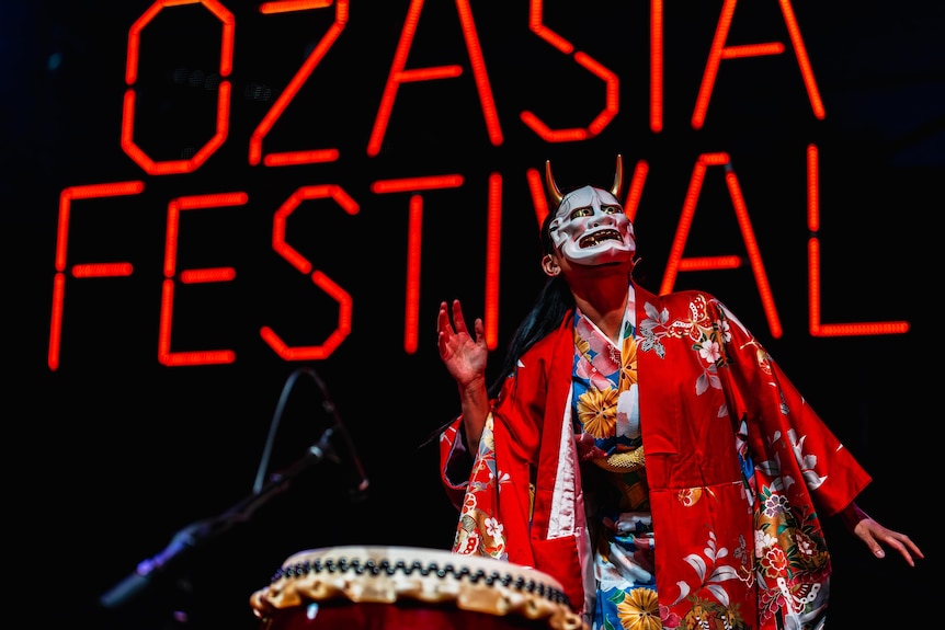 A female performer in a red kimono and a traditional face mask performs on stage in front of a red OzAsia Festival sign