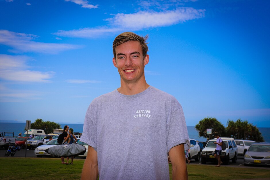 A young man wearing a grey shirt standing in front of a parking lot with the ocean visible in the background.