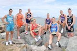 Eight women in their basketball gear, sold holding basketballs, sit or stand on rocks in front of water and smile at the camera.