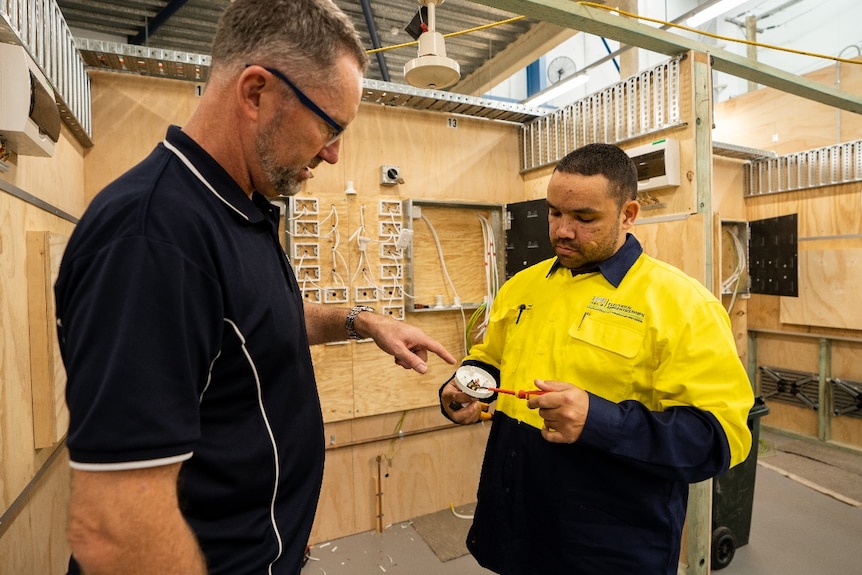 A young man wearing a high-vis shirt tinkers with an electrical part while an older man points to a piece inside it.