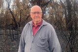 A man with a serious look on his face stands in front of burnt bushland.