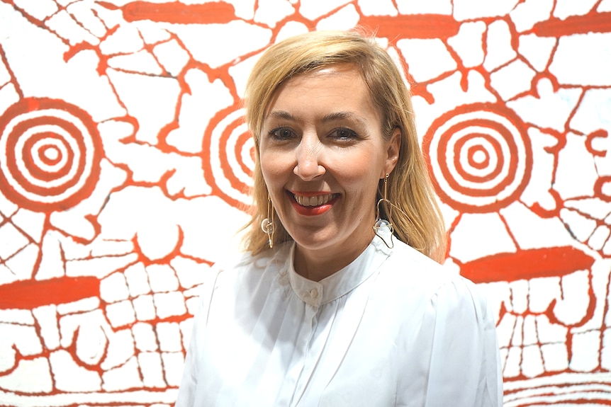 A blonde woman stands smiling in front an Aboriginal painting