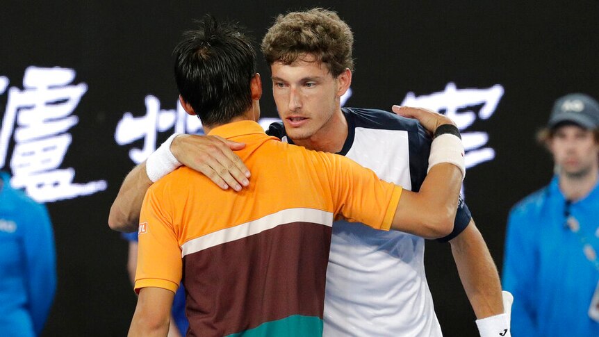 Two tennis players, one wearing an orange, brown and green shirt, and the other in blue and white, hug each other