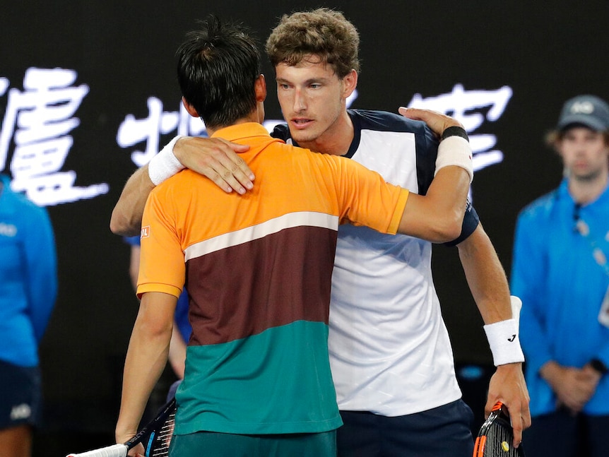 Two tennis players, one wearing an orange, brown and green shirt, and the other in blue and white, hug each other