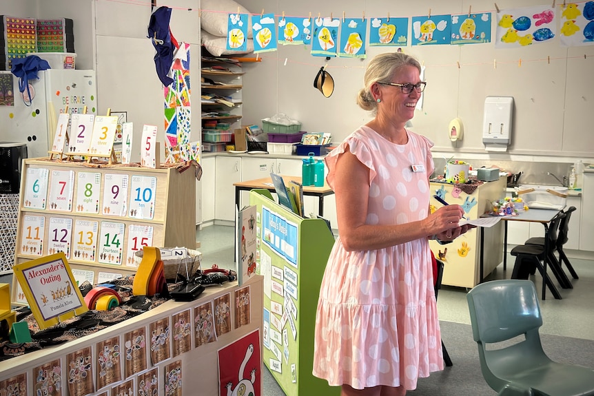 A woman holds a writing pad while standing in a kindergarten classroom surrounded by children's art