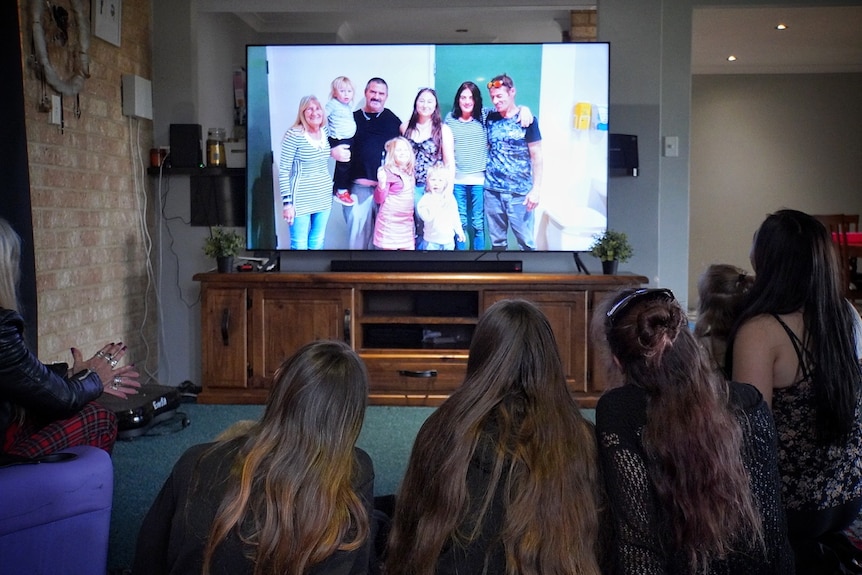 A family pictured from behind as they sit at a lounge room looking at a family photo on a TV