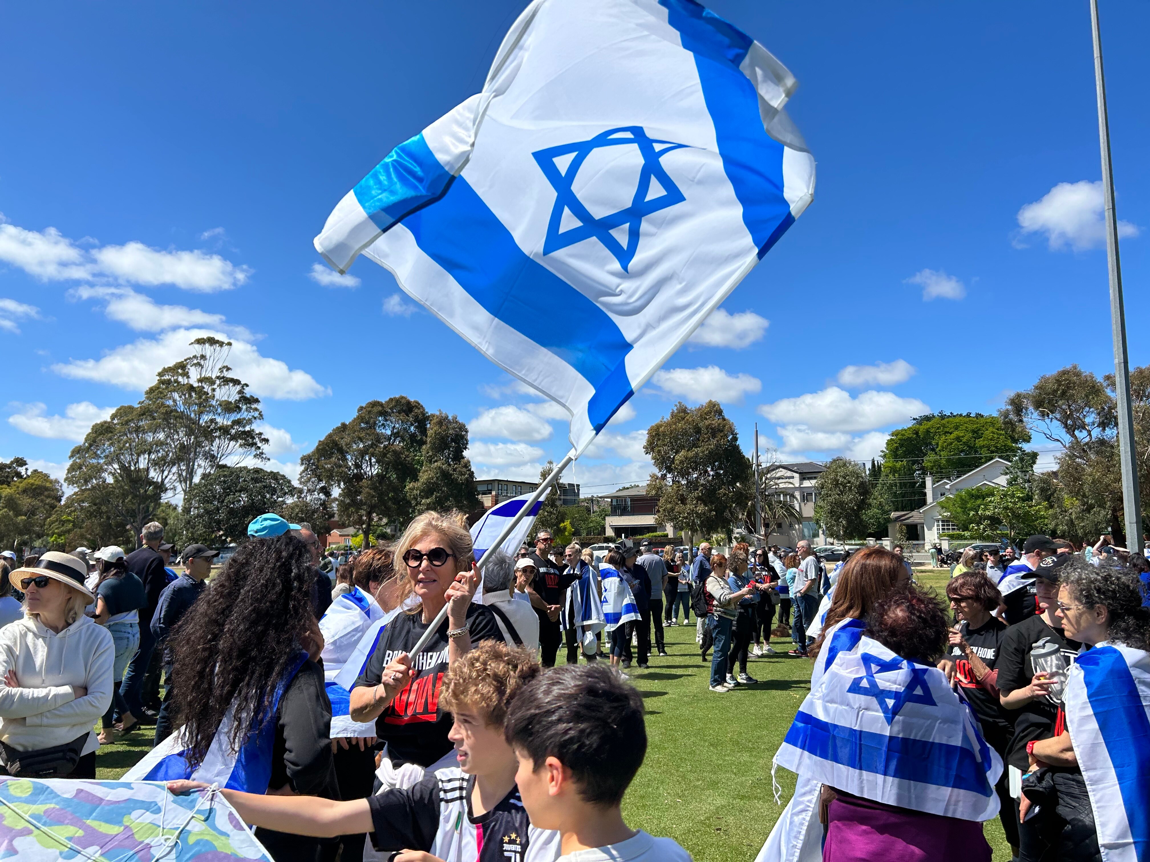 Members of a crowd of about 200 people fly Israeli flags in a park on a sunny day.