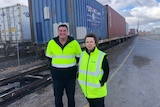 A man and a lady wearing hi vis jackets stand beside a train that's being loaded and unloaded