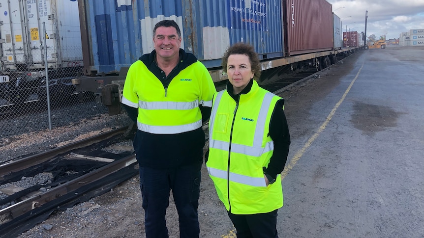 A man and a lady wearing hi vis jackets stand beside a train that's being loaded and unloaded