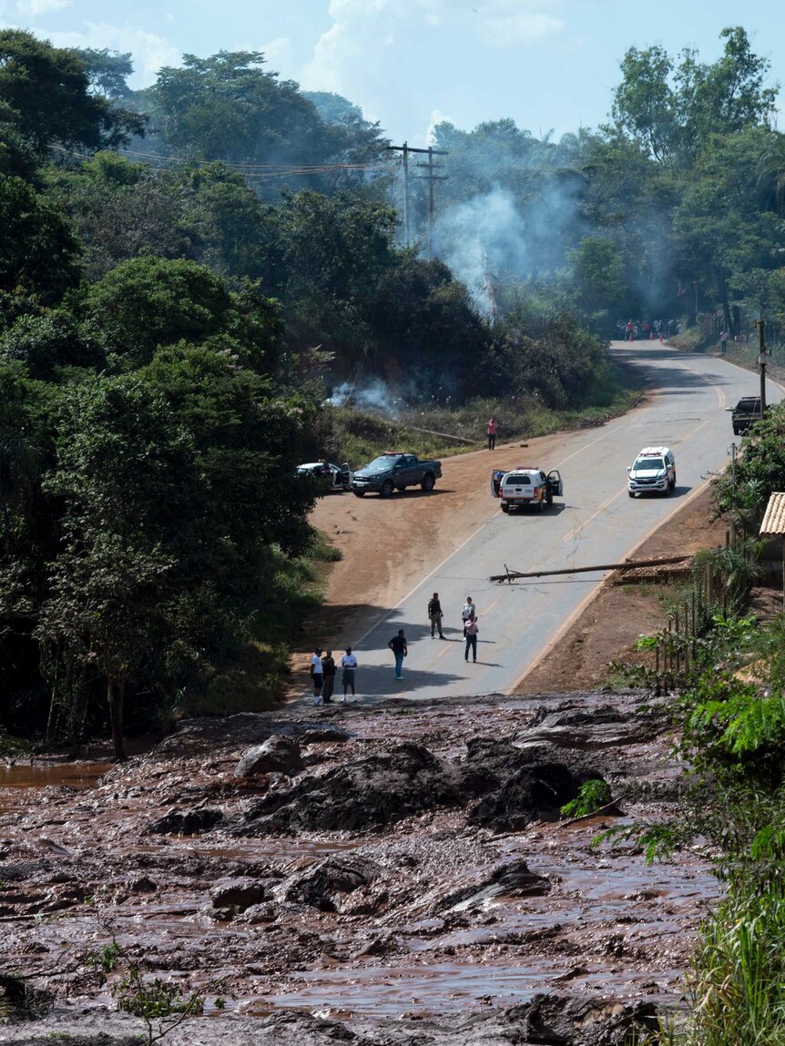 A two-lane road winding through forest is blocked by thick brown sludge. Smoke wafts through trees and cars park on the road.