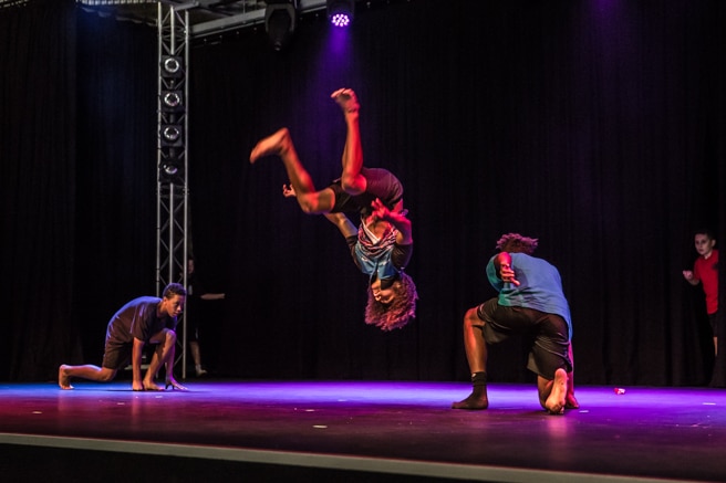 A dancer jumps upside down on a stage with two other dancers watching on