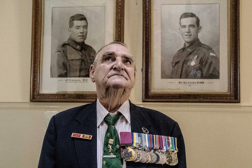 A man in his eighties, wearing his medals, standing in front of black-and-white framed photos of servicemen