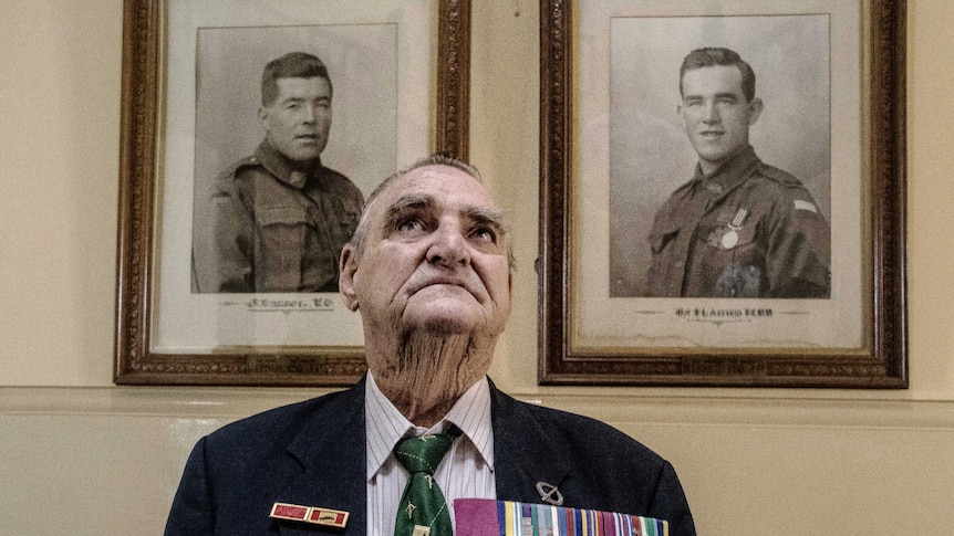 A man in his eighties, wearing his medals, standing in front of black-and-white framed photos of servicemen