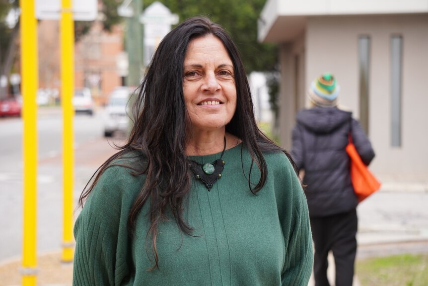 Sharon wears a pastel dark green jumper, standing on a street corner looking into the camera smiling