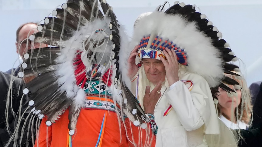 Pope Francis wears a traditional head dress and stands facing another person in traditional dress. 