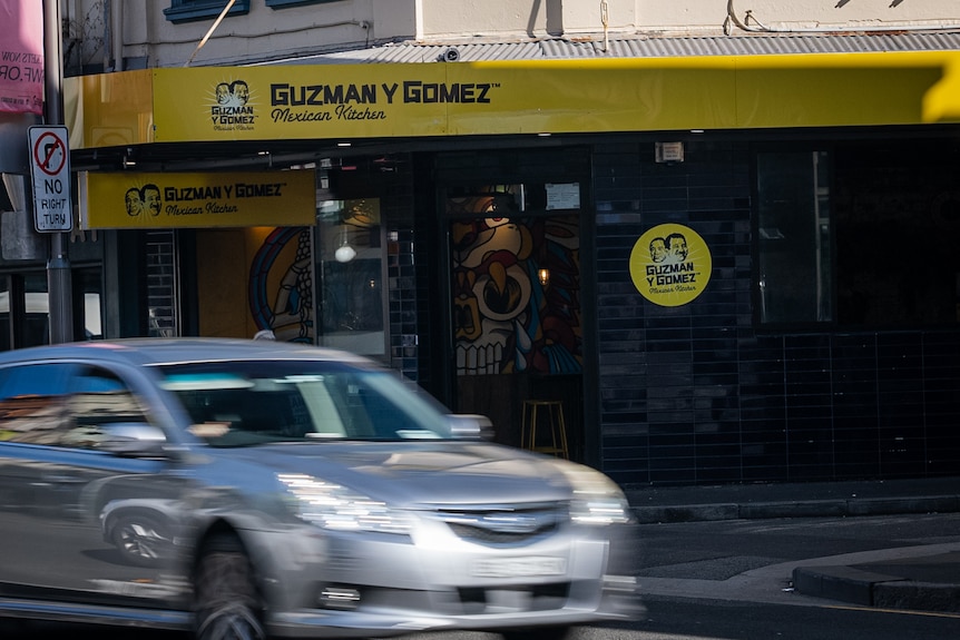 A view of a fast-food restaurant with yellow trim wrapping the building in its logo. A silver car is seen driving past.