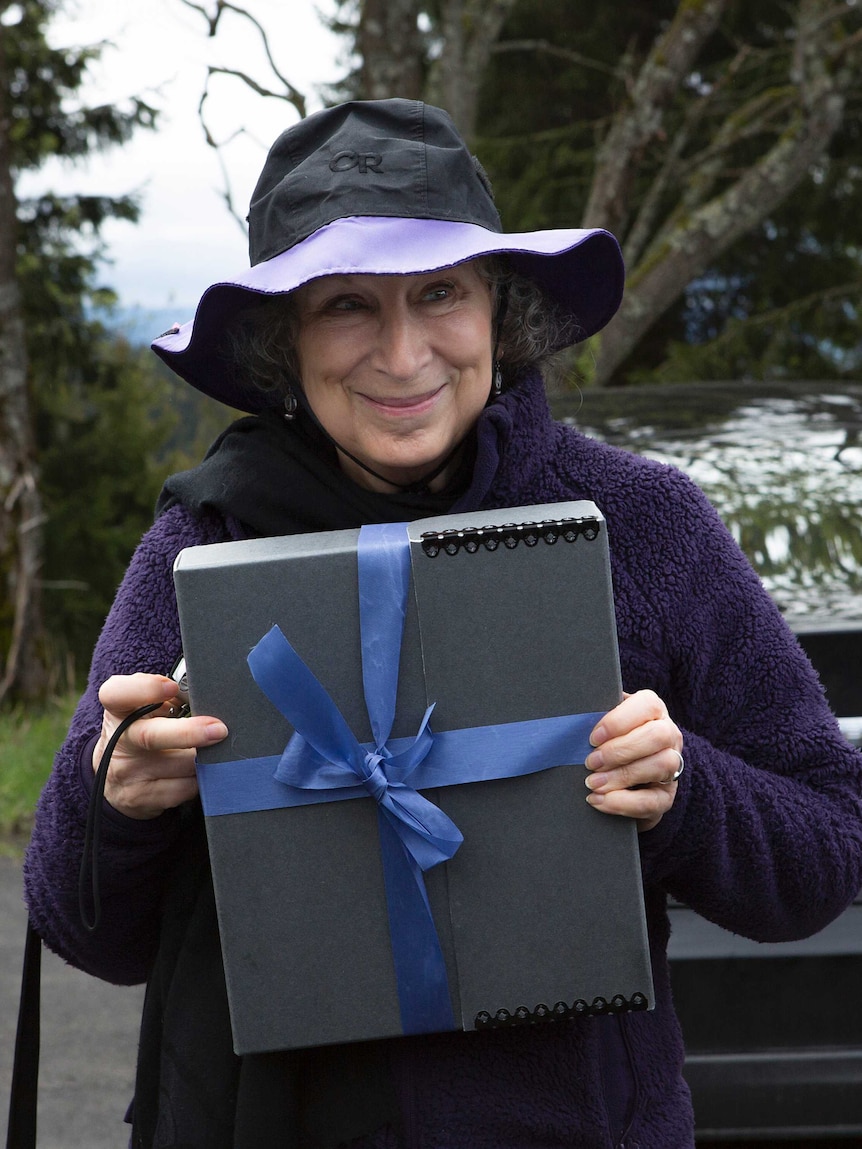 A woman in a purple jacket and a black and purple hat stands smiling at the camera, holding a grey box wrapped in a blue ribbon.