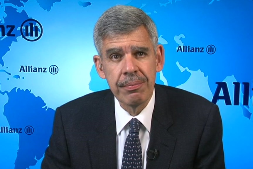 Close up shot of a man in a black suit looking directly at camera, with a blue background