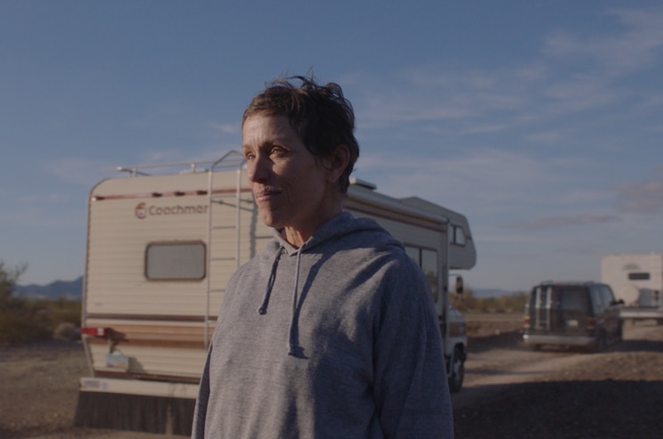 Actress with pixie haircut and no make-up, wearing grey hoodie, with campervan and country road on flat landscape in background.