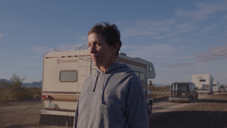 Actress with pixie haircut and no make-up, wearing grey hoodie, with campervan and country road on flat landscape in background.