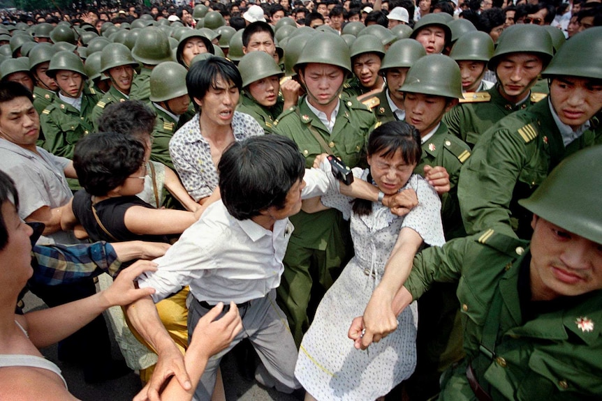 A young woman is vastly outnumbered by Chinese soldiers in green military uniform as they wrest her from other civilians.