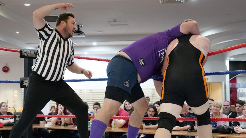 A referee stands over two men in a wrestling ring, while one of the men tried to choke the other as a crowd watches.