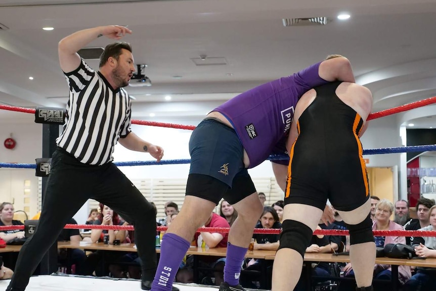 A referee stands over two men in a wrestling ring, while one of the men tries to choke the other as a crowd watches.