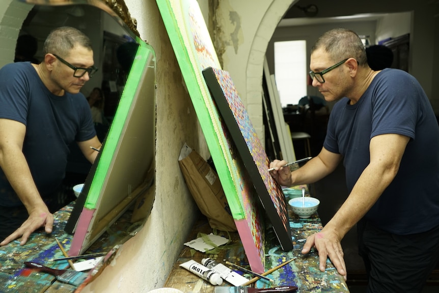 Daniel paints a painting on an easel which is reflected back by a mirror 