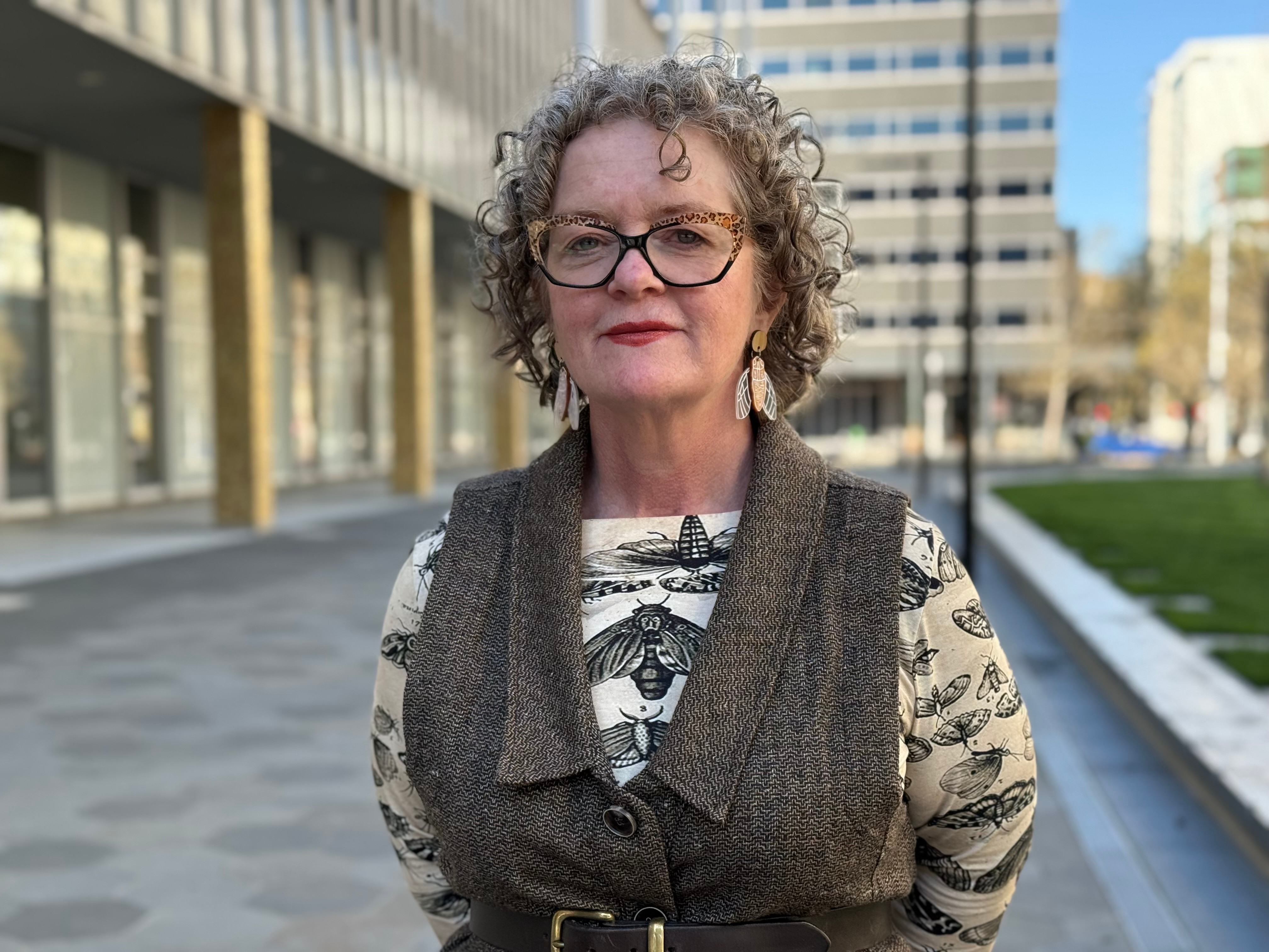 A woman with curly short hair, red lipstick and glasses wearing an insect-print top with insect earrings.