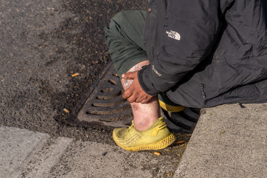 A man with yellow sneakers sitting on the concrete next to a gutter.