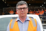 A man stands in high-visibility jacket in front of a group of apprentice in the distant background. 
