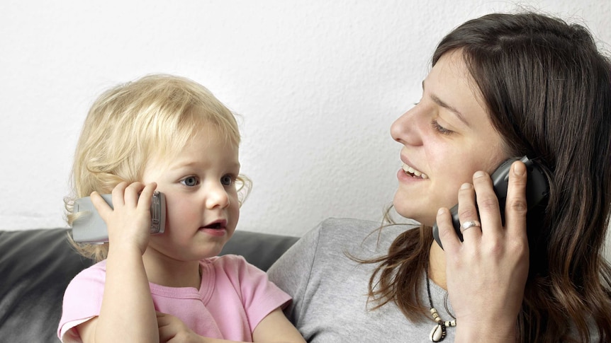 A toddler holds up a mobile phone to her ear, mirroring her mum who is doing the same