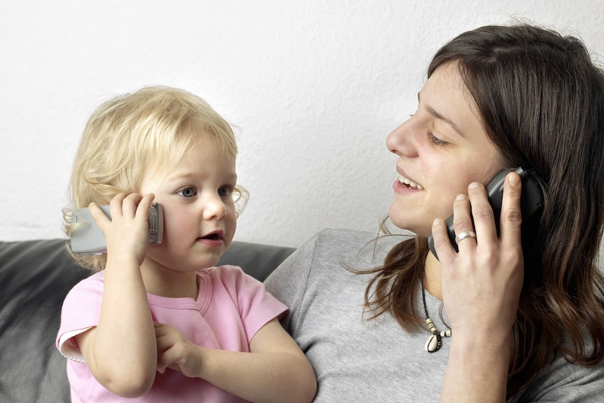 A toddler holds up a mobile phone to her ear, mirroring her mum who is doing the same
