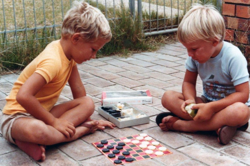 Two young boys with blonde hair sit on pavers outside playing checkers