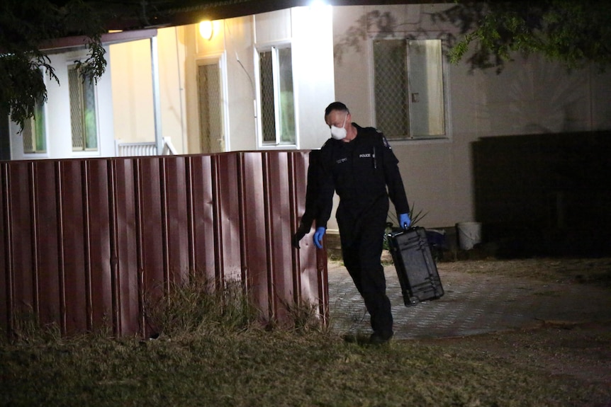 A police forensic officer walks out of Cleo Smith's house at night and around a fence carrying a black case.