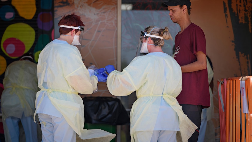 An Aboriginal man stands as two health workers in protective equipment hand a swab between them.