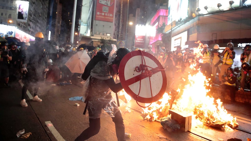 A protestor uses a shield to cover himself as he faces policemen while fire burns in the background