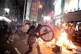 A protestor uses a shield to cover himself as he faces policemen while fire burns in the background