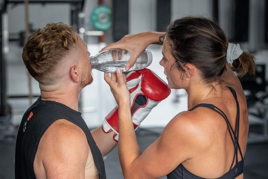 A young woman wearing a sports bra holds a bottle of water up for Luke to drink from. They have their backs to the camera.