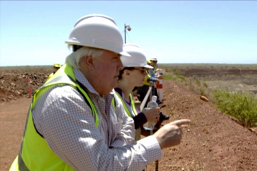Clive Palmer, wearing a high-visibility vest and hard hat, gestures towards a mine