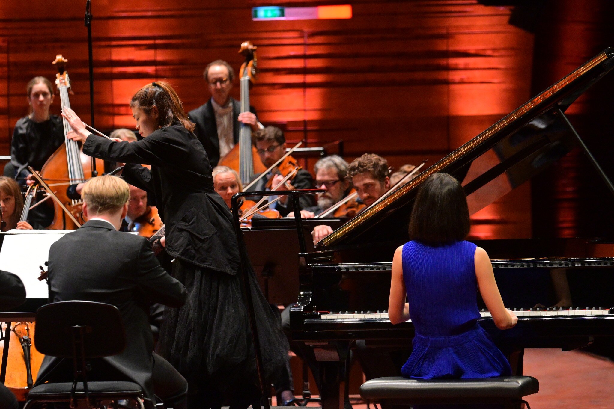 Pictured from behind, in a sleaveless bright blue dress, pianist Alice Sara Ott performs with orchestra.