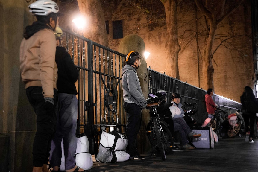 Cyclists in helmets with their bikes and bags lean against a fence outside a building.