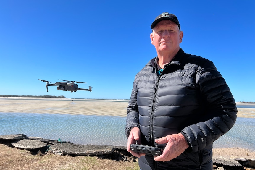 Man standing beside hovering drone, with beach in background